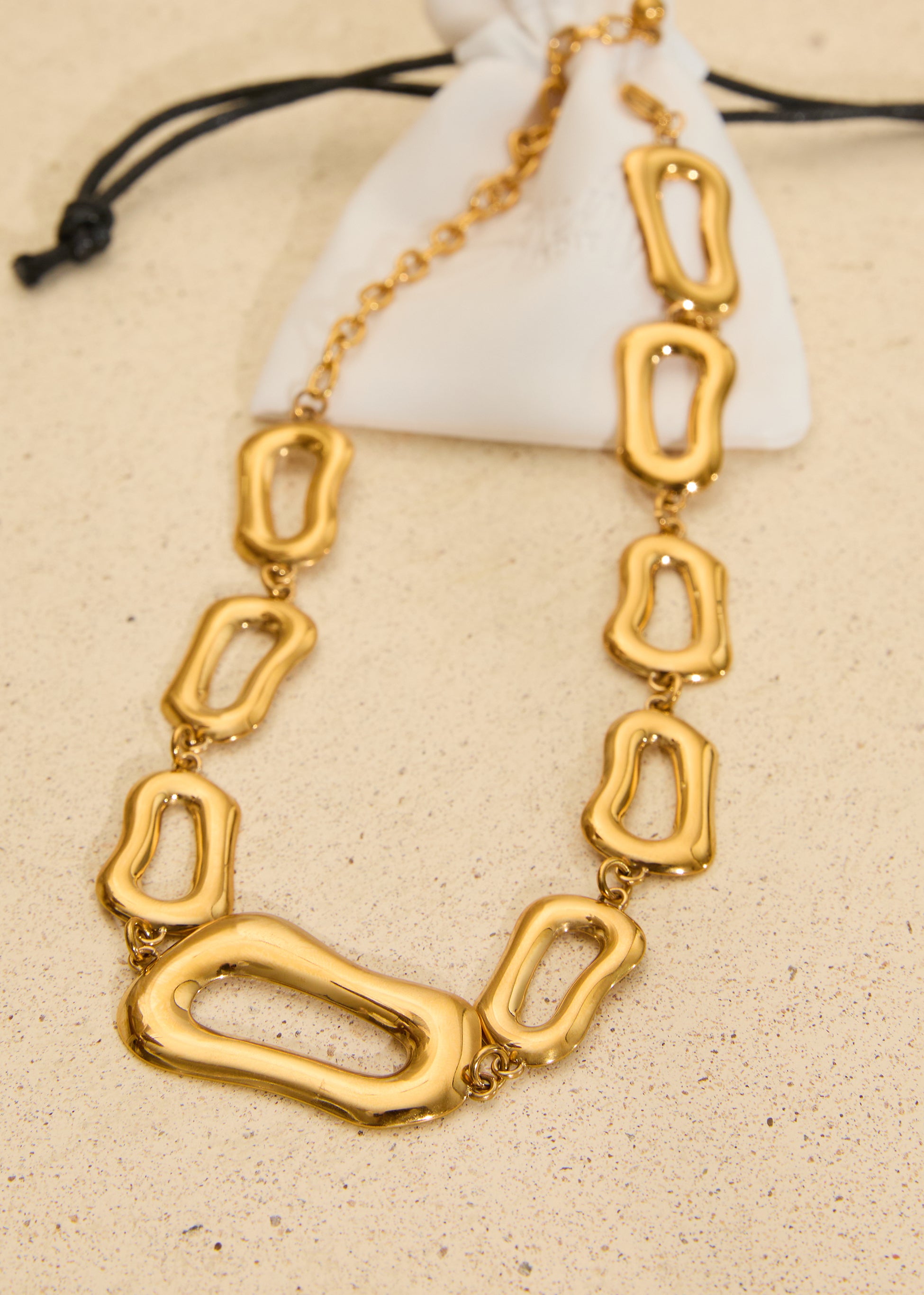 Gold stainless steel statement necklace featuring polished organic-shaped links displayed on a neutral background with a white jewellery pouch.