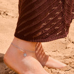 Silver layered evil eye anklet with a crystal tennis chain and paperclip link chain, featuring a round turquoise and crystal evil eye pendant, displayed on a The Seventh Edit pouch in sand.​​​​​​​​​​​​​​​​Uk jewellery 