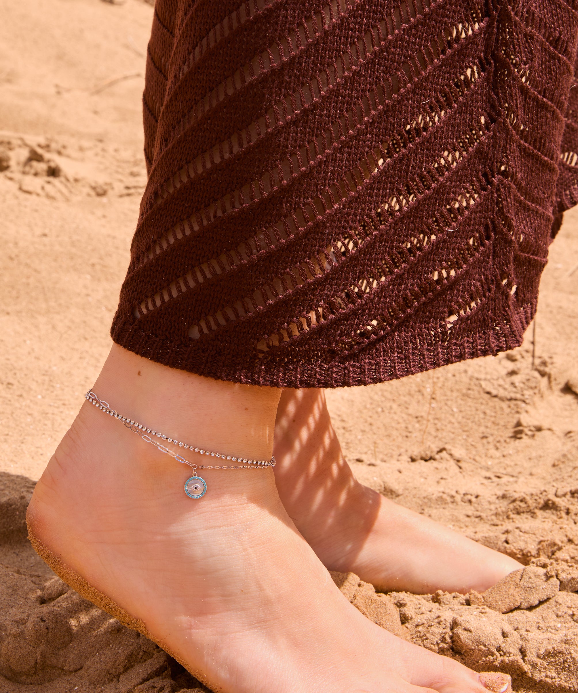 Silver layered evil eye anklet with a crystal tennis chain and paperclip link chain, featuring a round turquoise and crystal evil eye pendant, displayed on a The Seventh Edit pouch in sand.​​​​​​​​​​​​​​​​Uk jewellery 