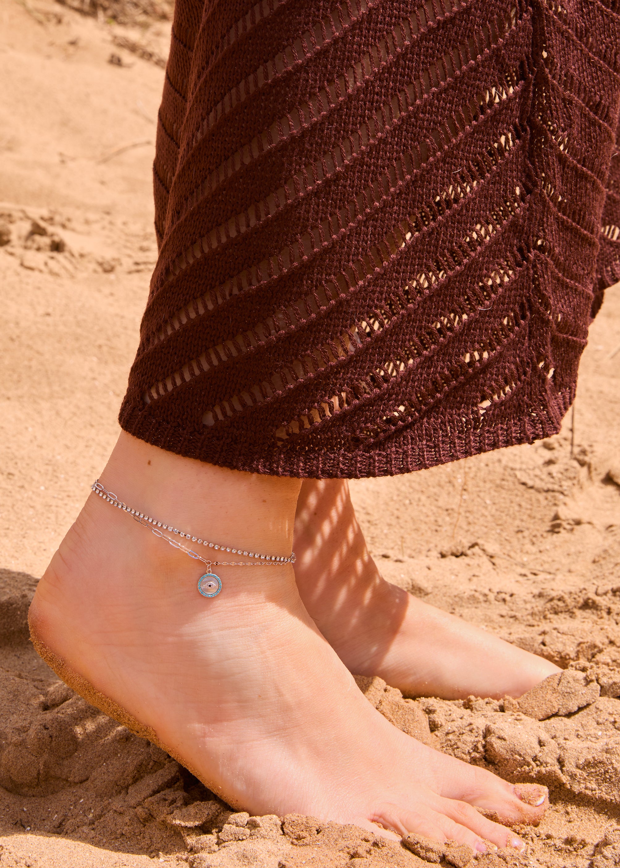 Silver layered evil eye anklet with a crystal tennis chain and paperclip link chain, featuring a round turquoise and crystal evil eye pendant, displayed on a The Seventh Edit pouch in sand.​​​​​​​​​​​​​​​​Uk jewellery 