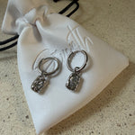 The Marlowe Hoops, gold hoop earrings with hammered charm and sparkling stone drop, laid on The Seventh Edit’s branded white pouch.