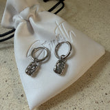 The Marlowe Hoops, gold hoop earrings with hammered charm and sparkling stone drop, laid on The Seventh Edit’s branded white pouch.