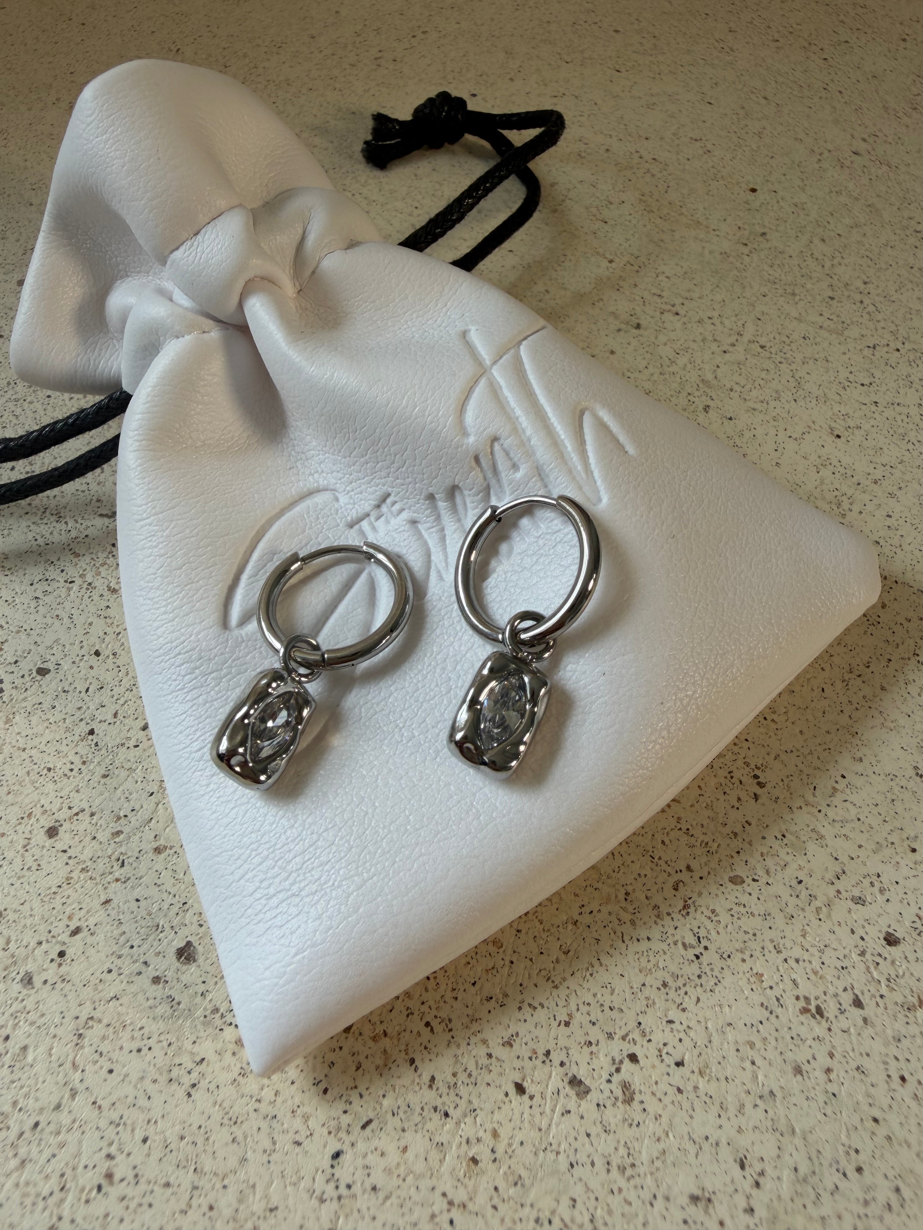 The Marlowe Hoops, gold hoop earrings with hammered charm and sparkling stone drop, laid on The Seventh Edit’s branded white pouch.