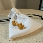 The Nico Hoops, gold ribbed huggie hoop earrings, laid on The Seventh Edit’s branded white pouch.