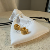 The Nico Hoops, gold ribbed huggie hoop earrings, laid on The Seventh Edit’s branded white pouch.