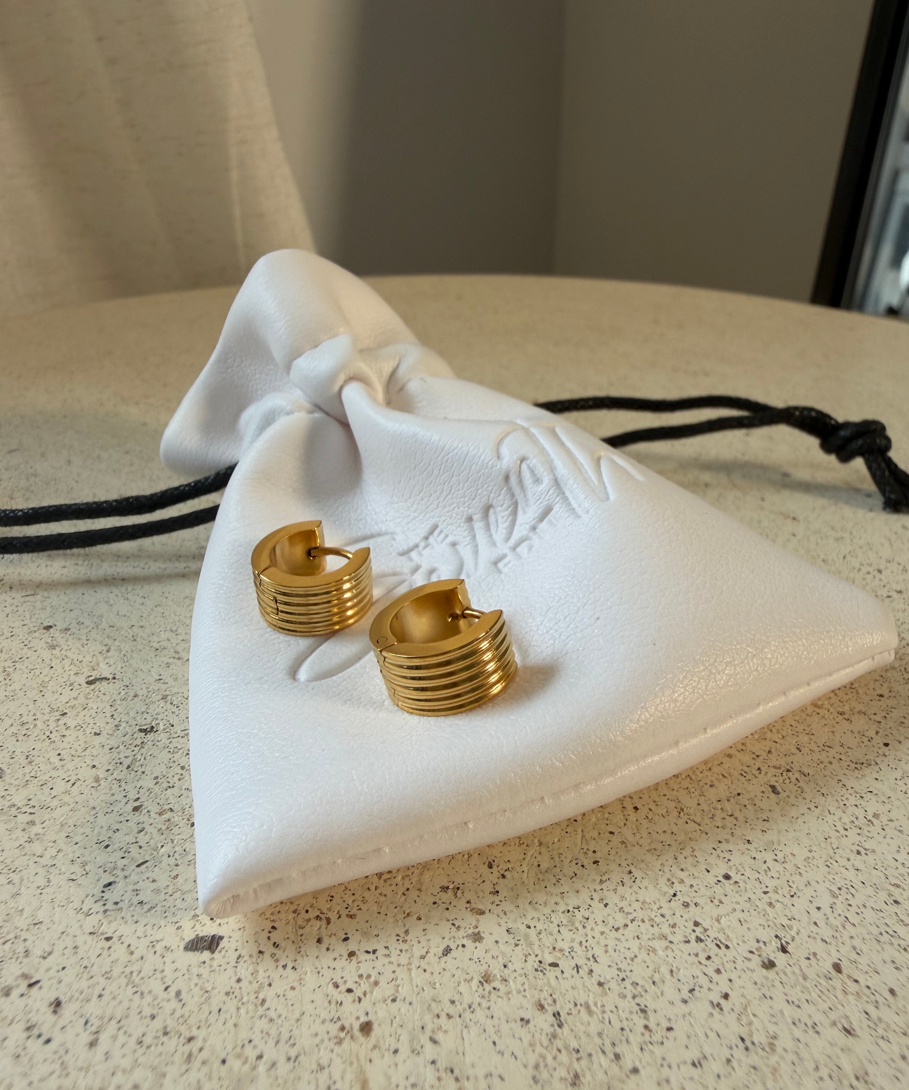 The Nico Hoops, gold ribbed huggie hoop earrings, laid on The Seventh Edit’s branded white pouch.