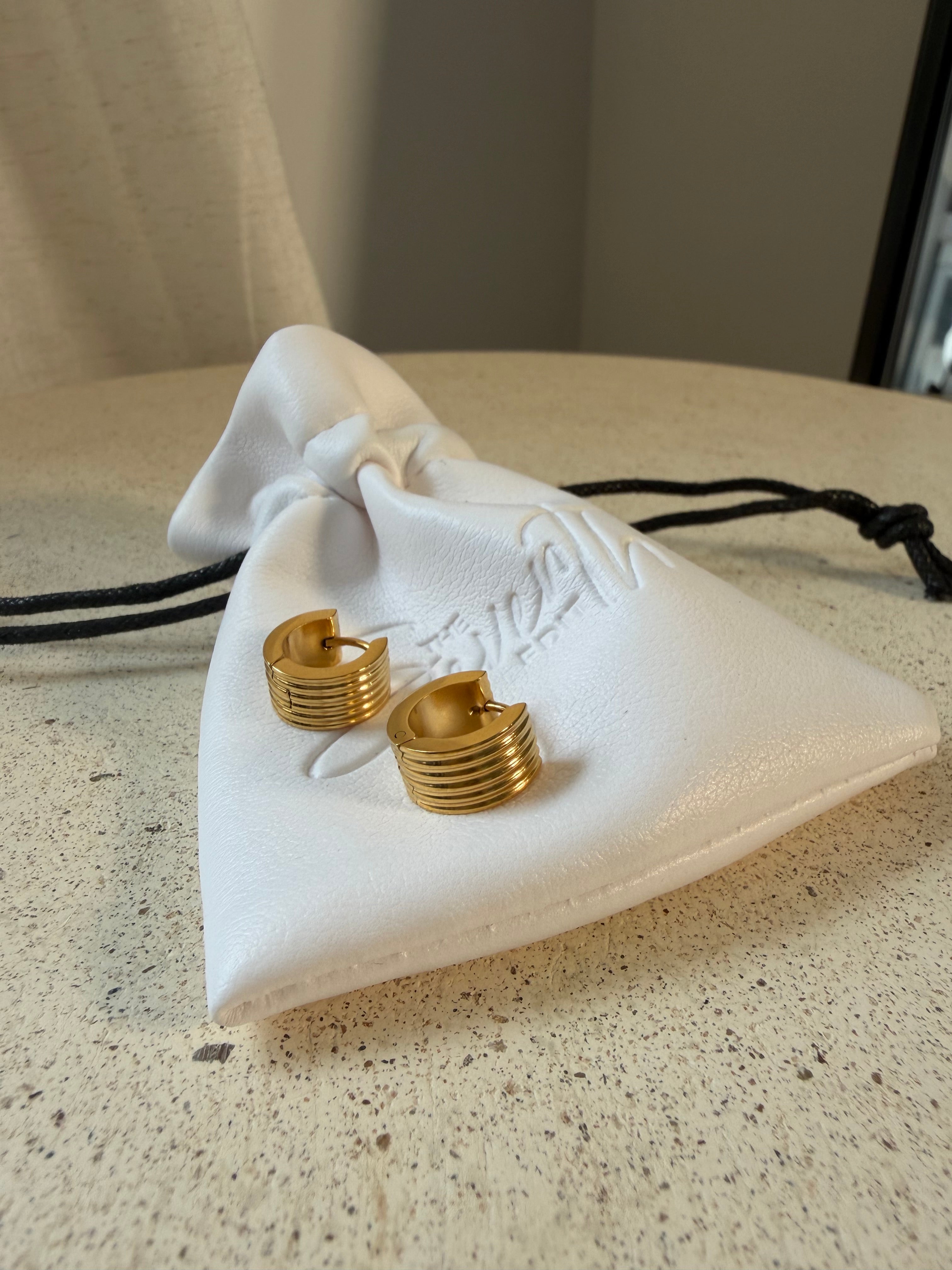 The Nico Hoops, gold ribbed huggie hoop earrings, laid on The Seventh Edit’s branded white pouch.