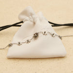 The Seventh Edit Daisy Anklet — silver daisy flower charm anklet laid across a white branded pouch on sand.