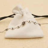 The Seventh Edit Daisy Anklet — silver daisy flower charm anklet laid across a white branded pouch on sand.