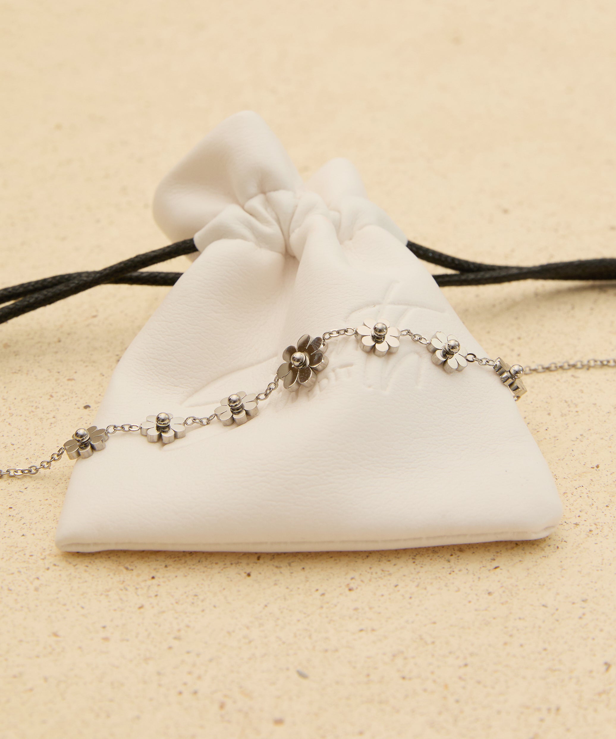 The Seventh Edit Daisy Anklet — silver daisy flower charm anklet laid across a white branded pouch on sand.