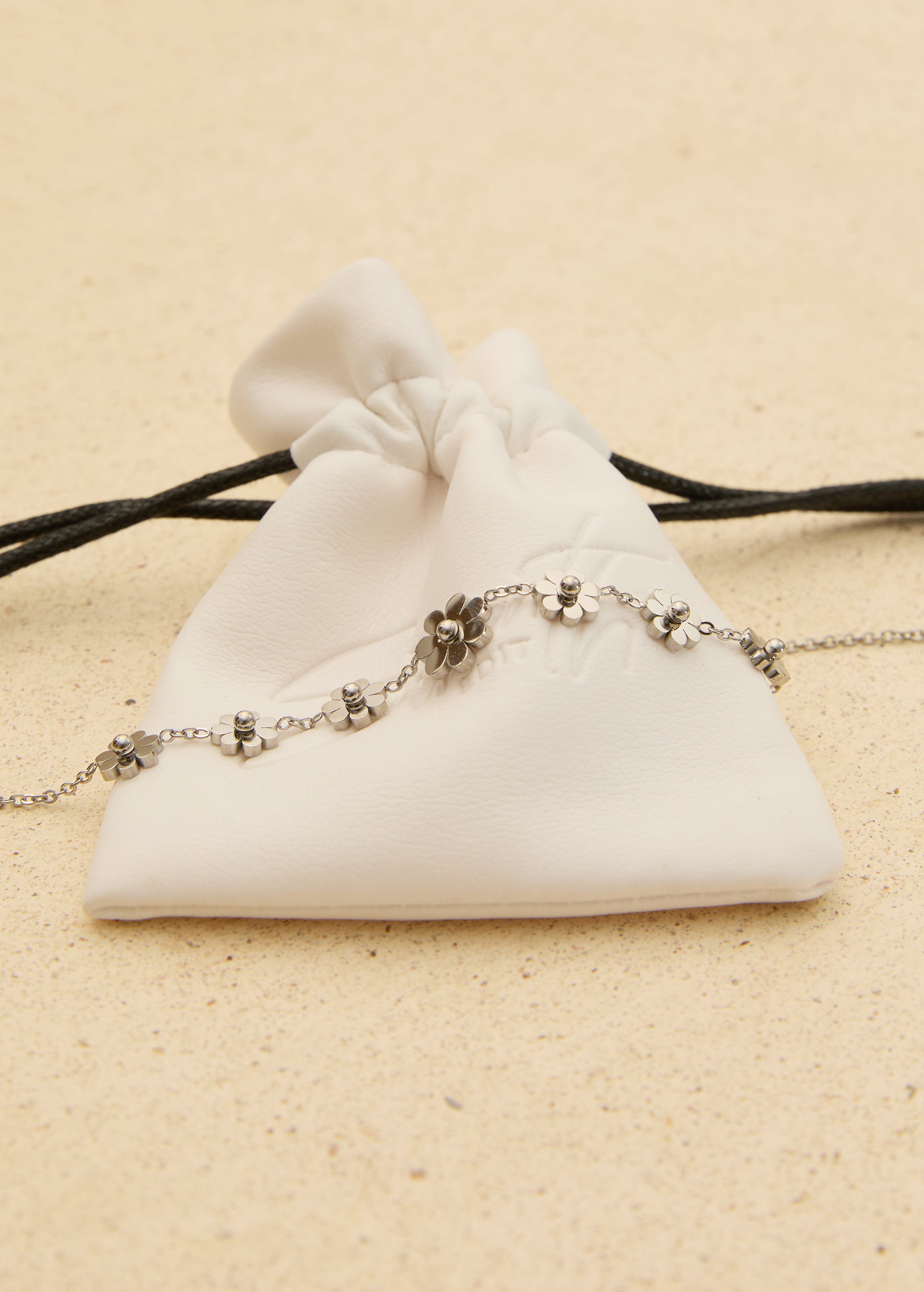 The Seventh Edit Daisy Anklet — silver daisy flower charm anklet laid across a white branded pouch on sand.