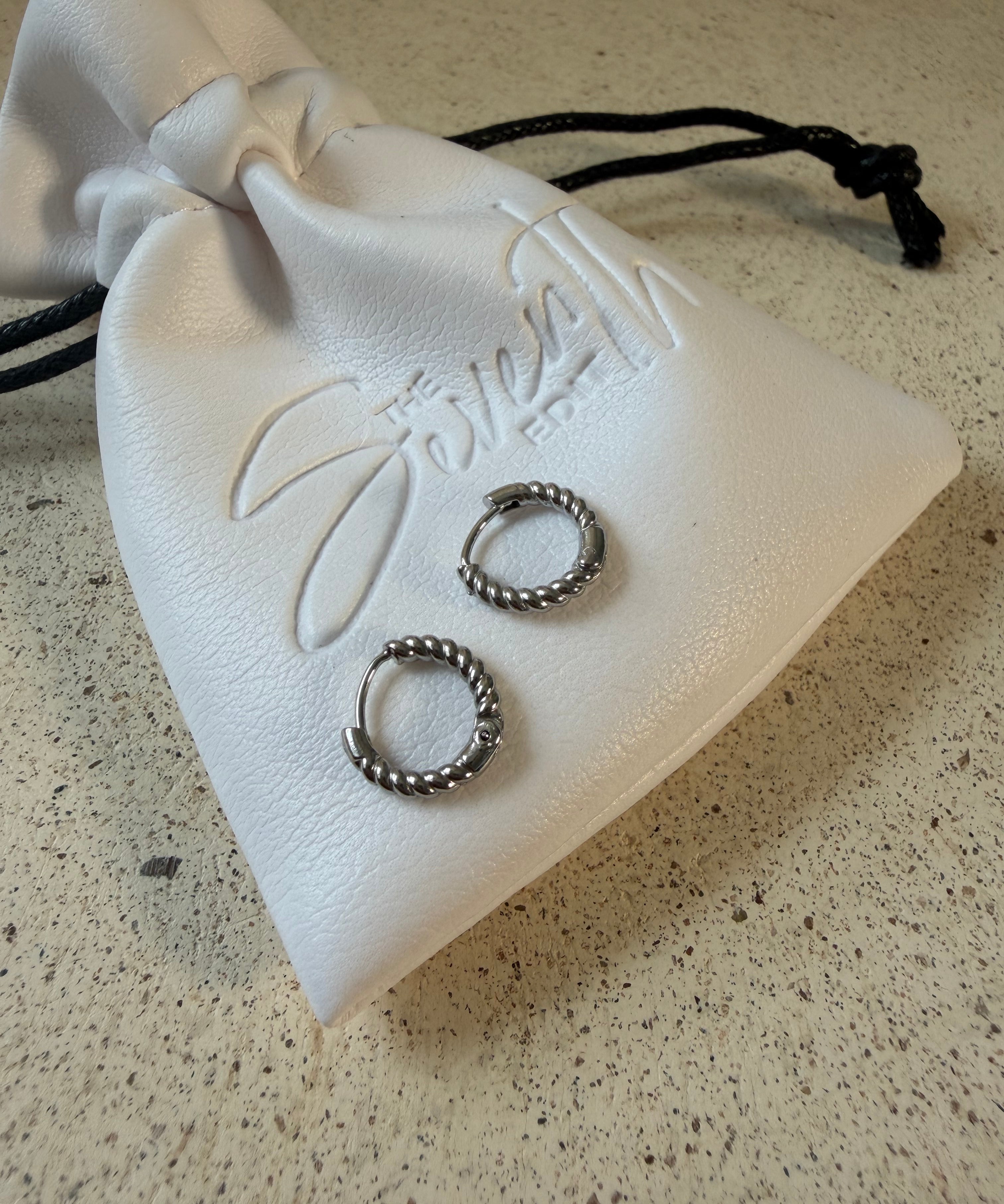 The Rio Hoops, silver twisted rope huggie hoop earrings, laid on The Seventh Edit’s branded white pouch.