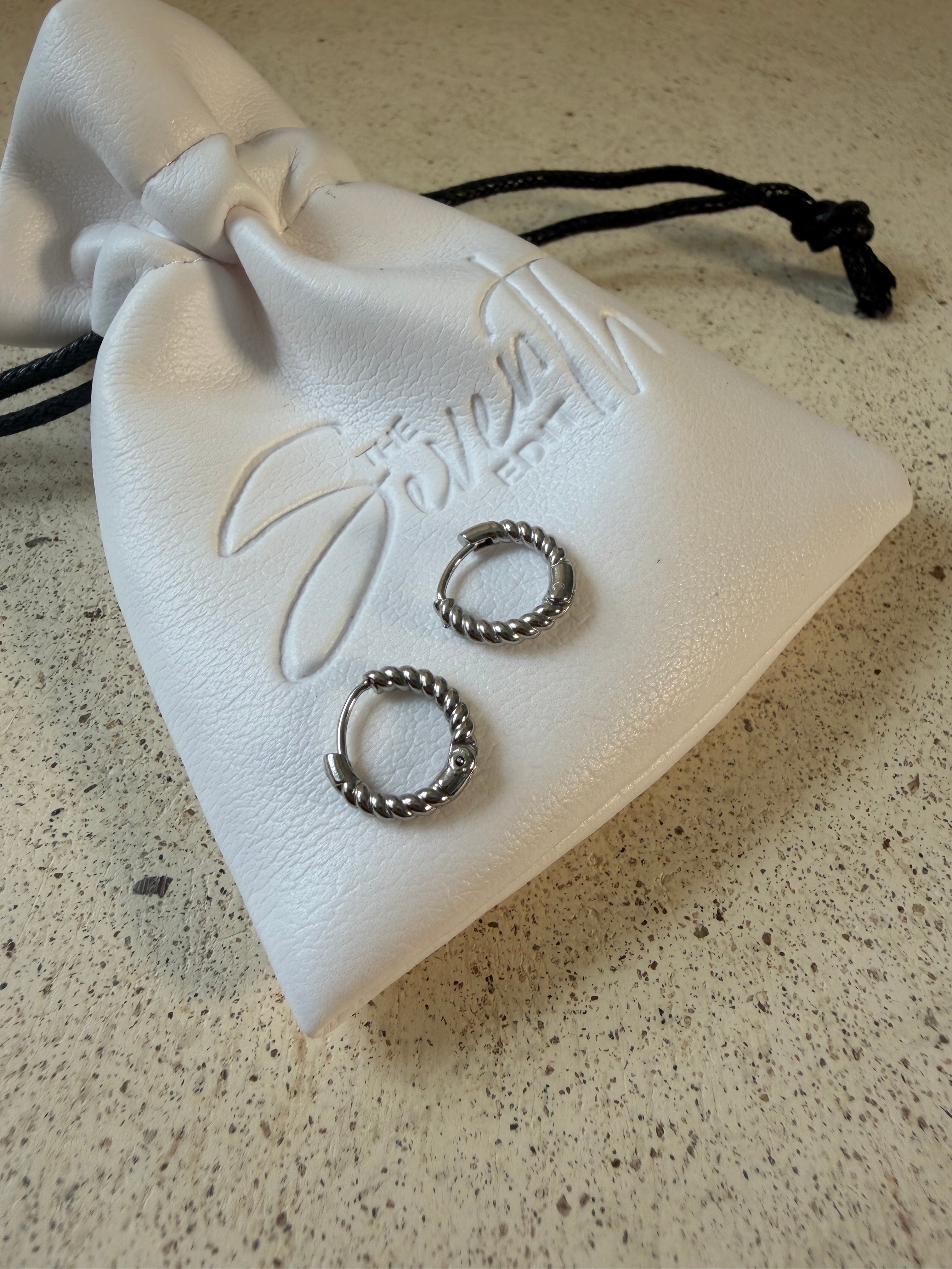 The Rio Hoops, silver twisted rope huggie hoop earrings, laid on The Seventh Edit’s branded white pouch.