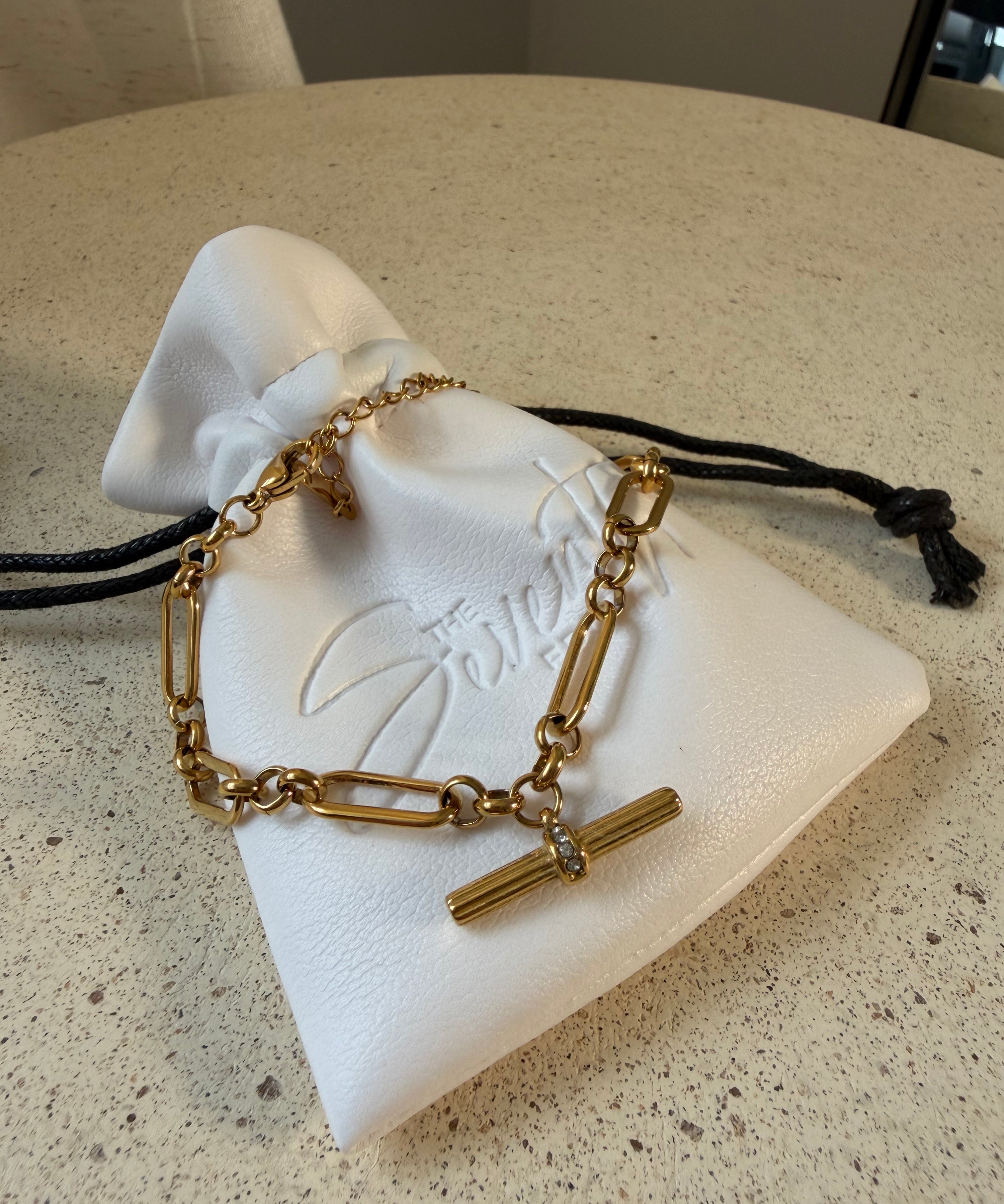 The Blake Bracelet, a waterproof gold paperclip chain bracelet with pavé T-bar toggle, laid on The Seventh Edit’s branded white pouch.