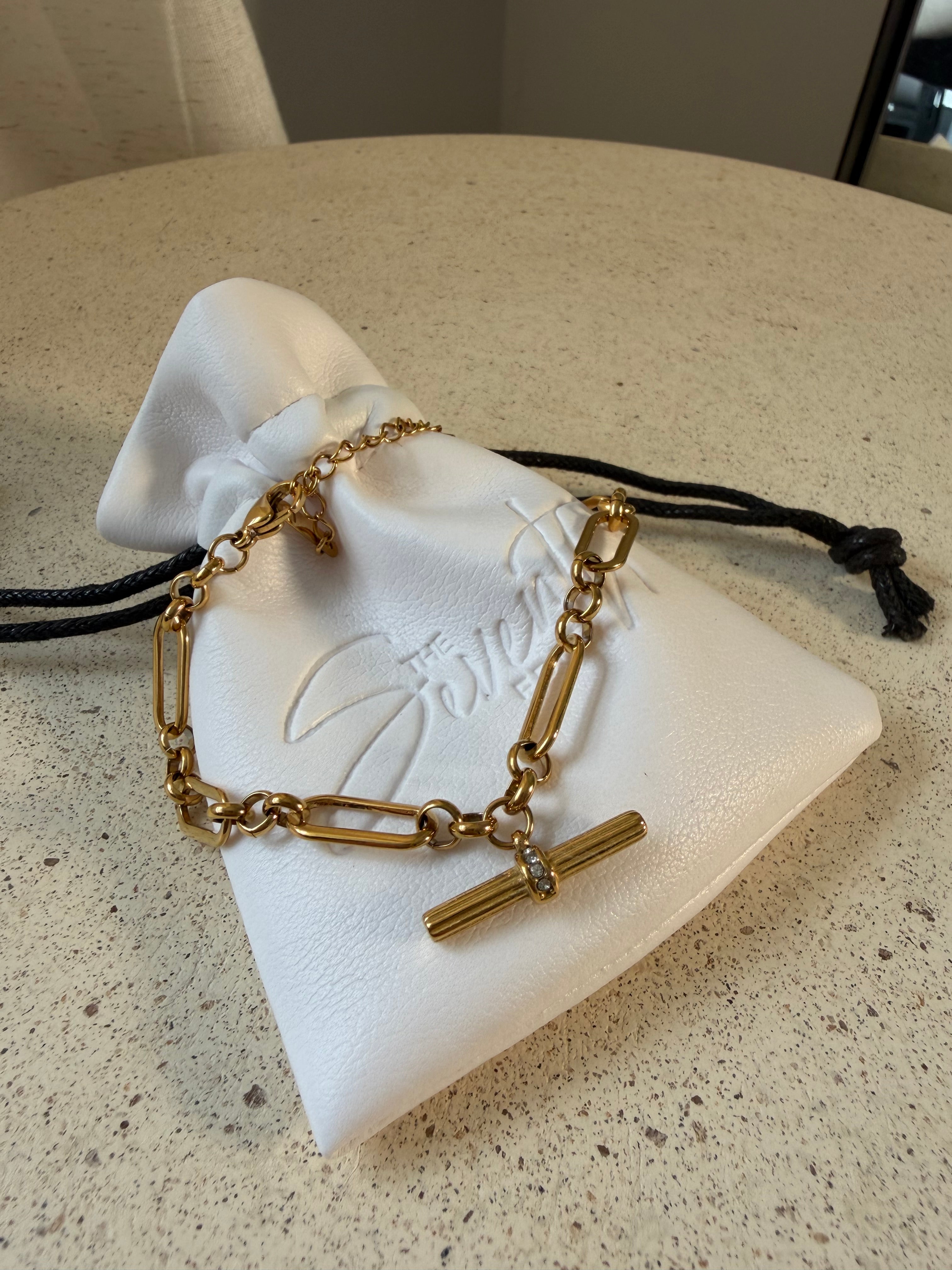 The Blake Bracelet, a waterproof gold paperclip chain bracelet with pavé T-bar toggle, laid on The Seventh Edit’s branded white pouch.