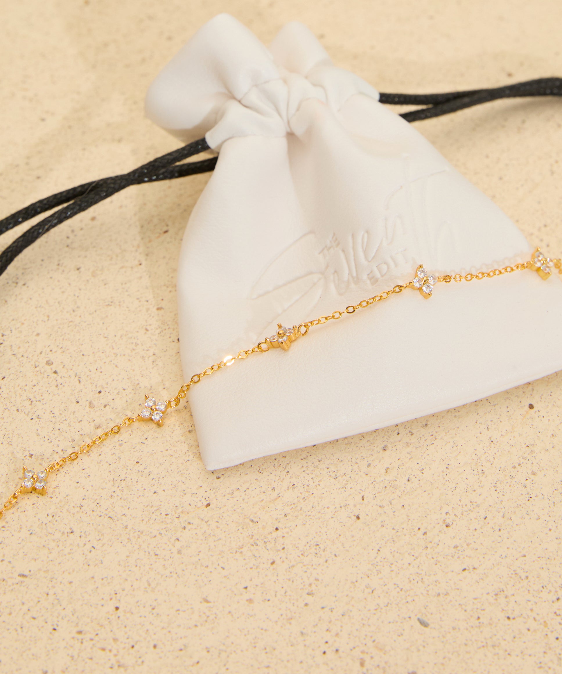 The Seventh Edit Stella Bracelet — gold crystal star charm chain bracelet draped across a white branded pouch on sand.
