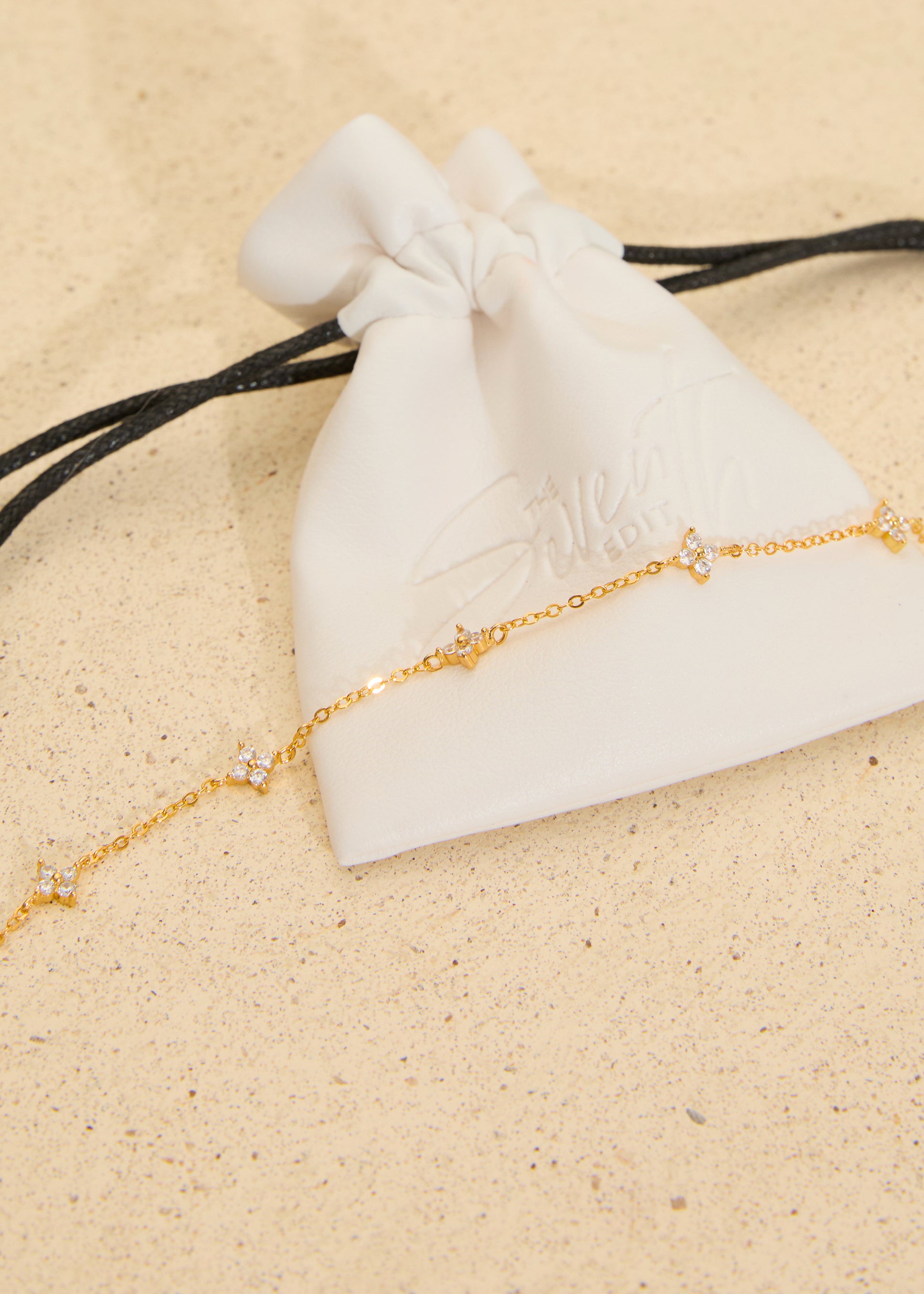 The Seventh Edit Stella Bracelet — gold crystal star charm chain bracelet draped across a white branded pouch on sand.