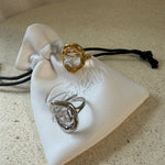 The Delphine Ring, a gold sculptural open ring with princess-cut stone, laid on The Seventh Edit’s branded white pouch.