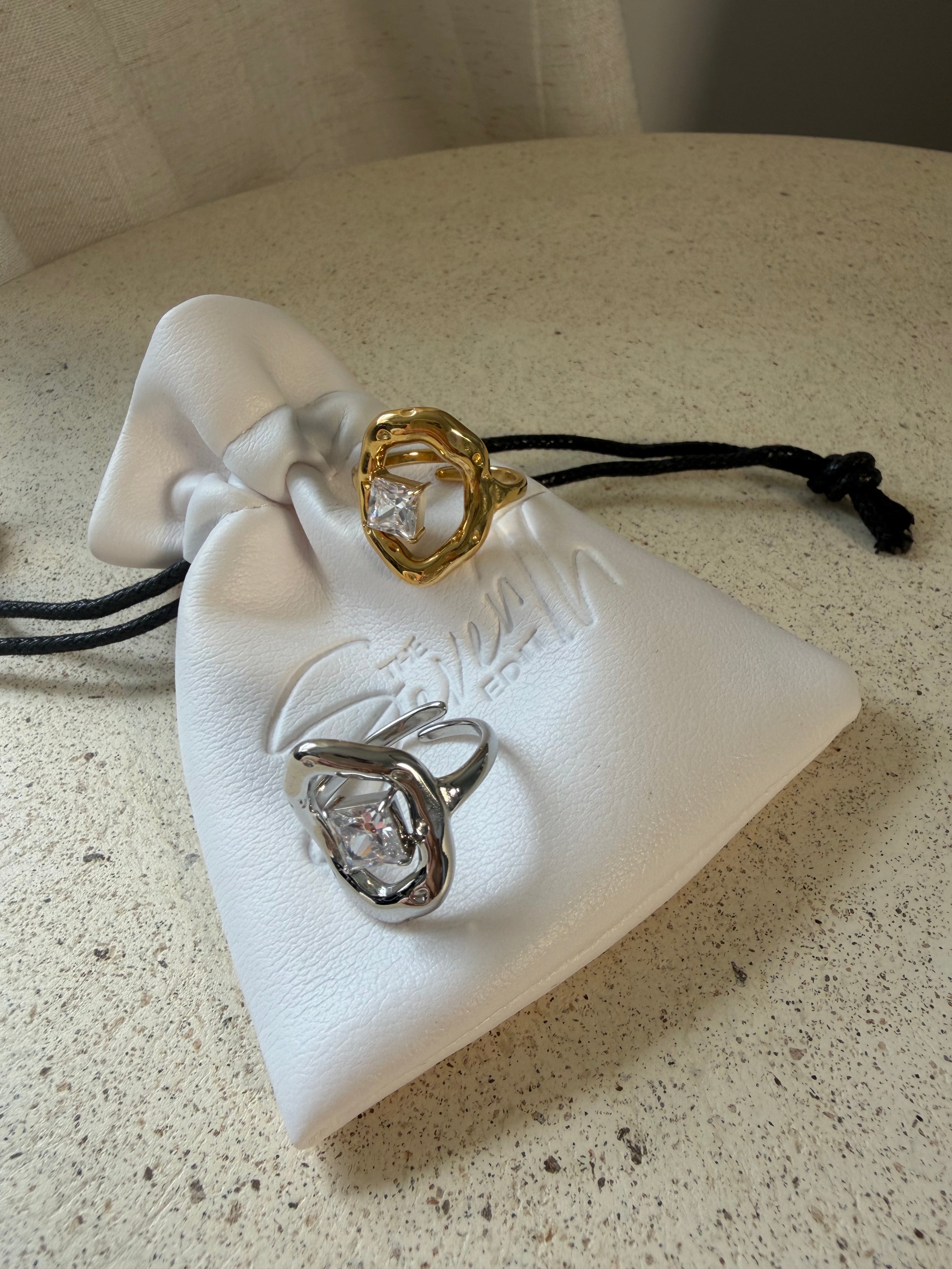 The Delphine Ring, a gold sculptural open ring with princess-cut stone, laid on The Seventh Edit’s branded white pouch.